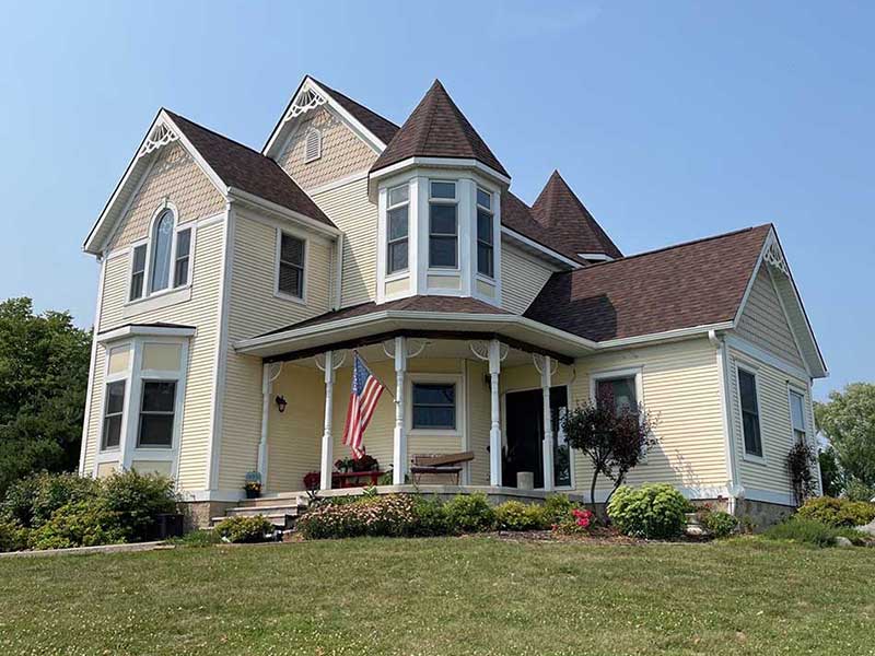 Front view of a large mid-class home with a front porch plus an American flag attached to one of its poles, and a red shingle roof worked on by American Dream Exteriors